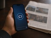 Una mano sostiene un smartphone en el que se ve el icono de un chatbot; junto a él hay un periódico impreso.
